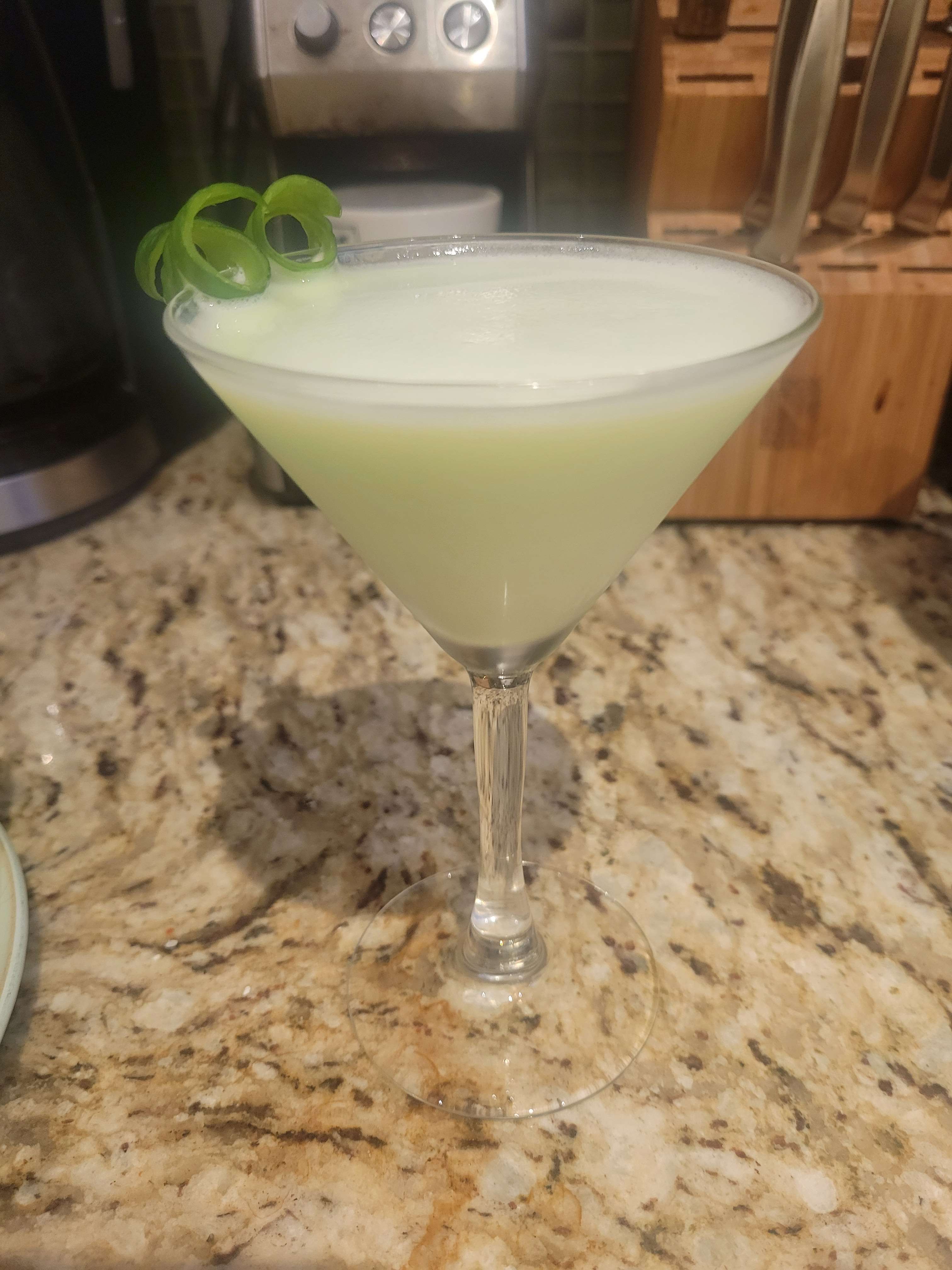 Image of the Macne Melon cocktail, a very light green opaque drink in a martini glass, garnished with two mini curled cucumber ribbons.